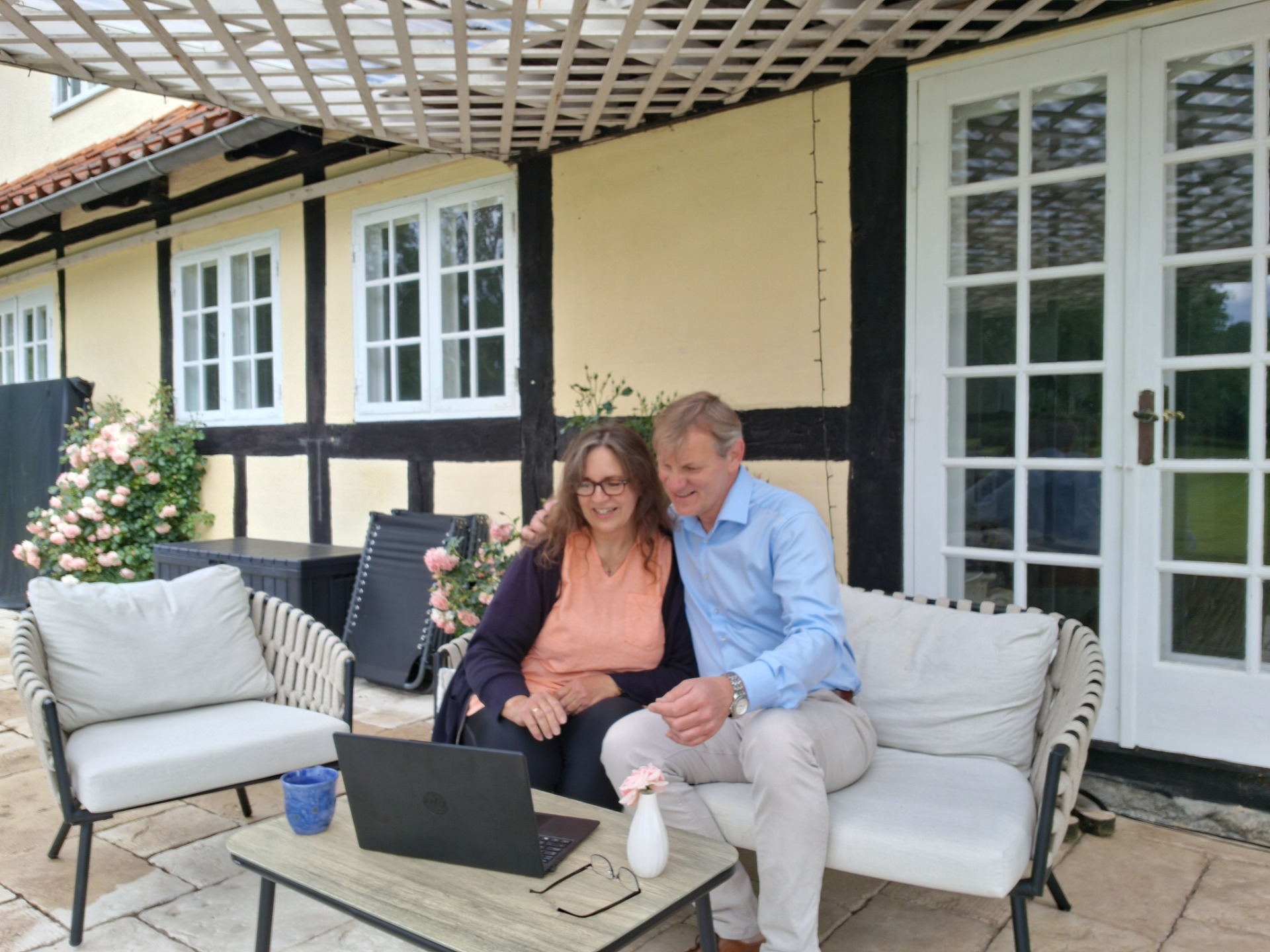 Par sidder i sofa på gårdhave foran hvidt bindingsværkshus, smiler og arbejder på laptop. Frihed og flexibelt arbejdsmiljø online.